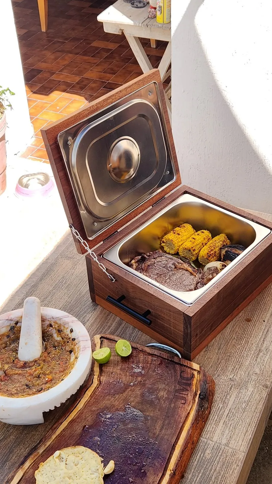 Grill Box con carne y elotes junto a un molcajete con salsa y una tabla de madera con limones