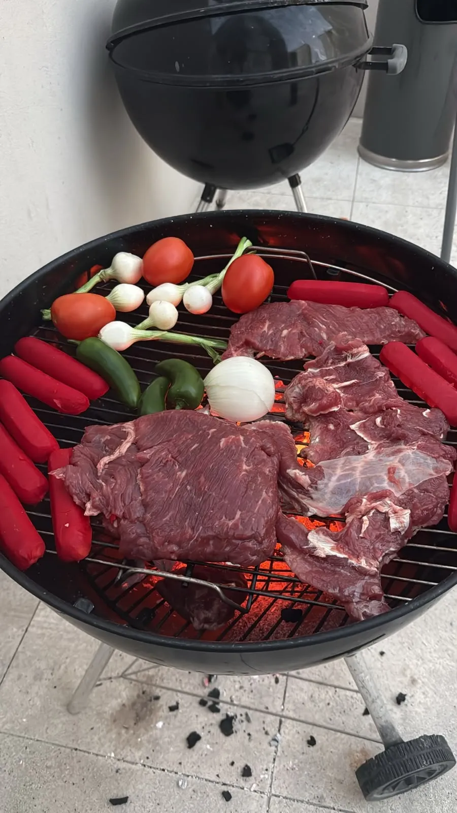 Asador de carbón con arrachera, salchichas, cebollitas, jitomates y chiles sobre las brasas