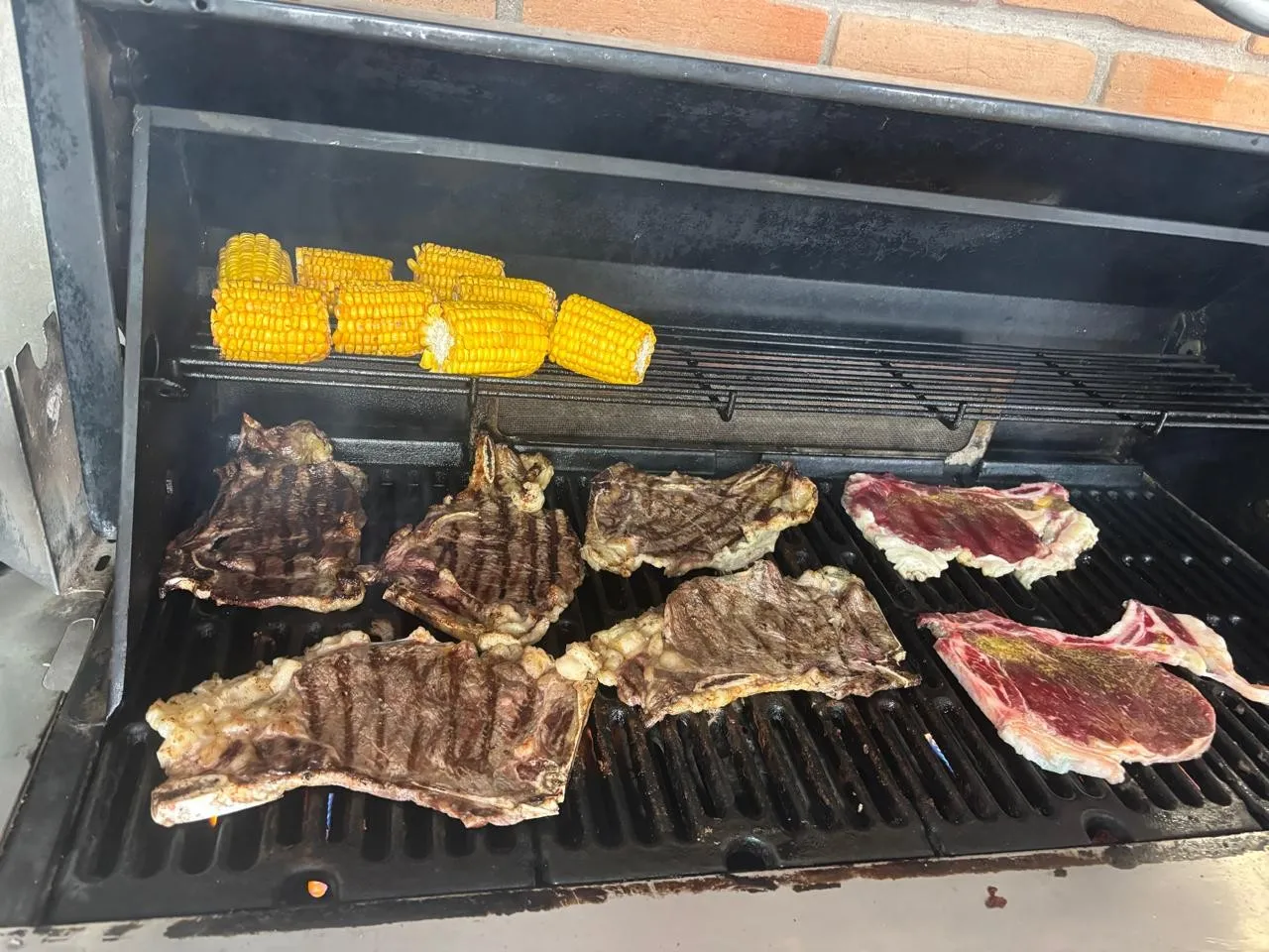 Cortes de carne asándose en la parrilla, con elotes en la rejilla de arriba