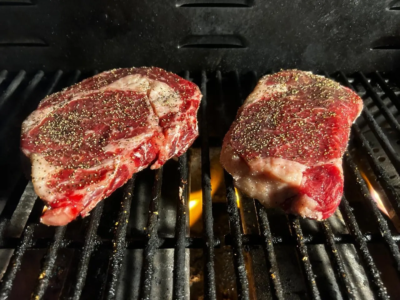 Un ribeye y un New York sazonados con pimienta sobre la parrilla, con la flama debajo