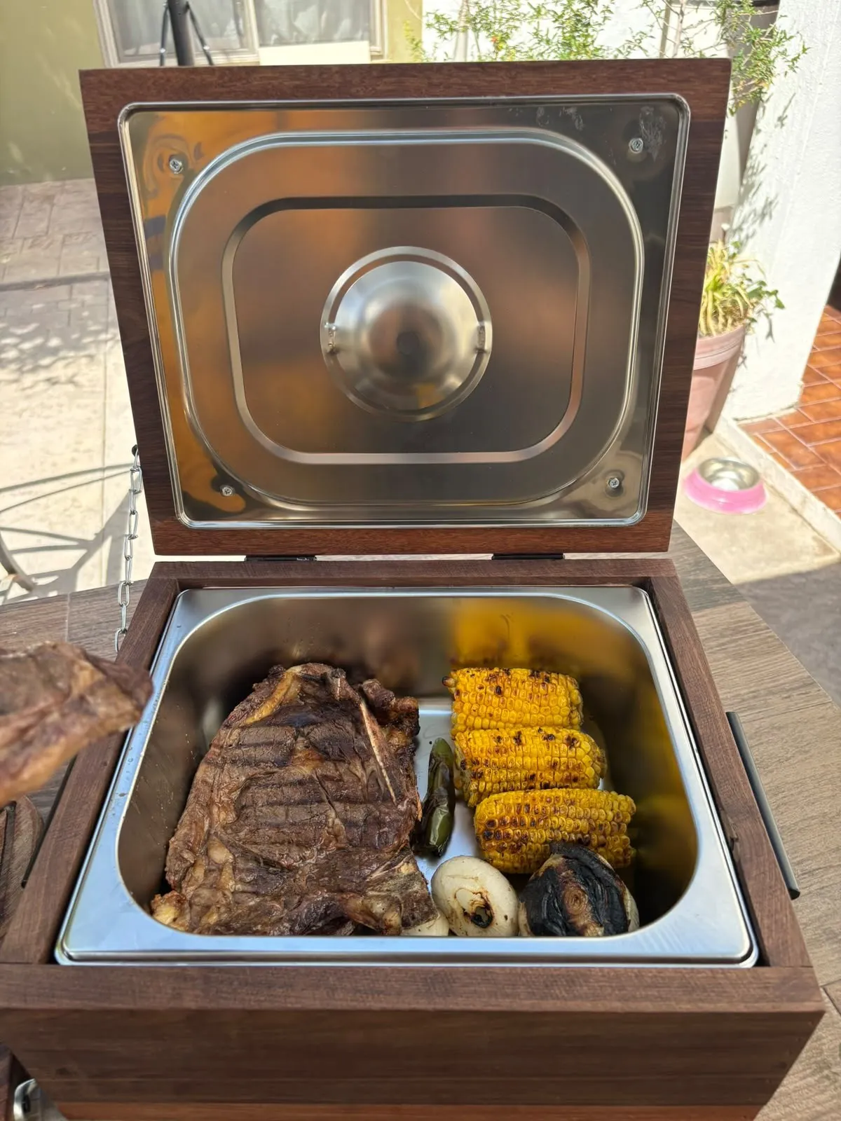 Grill Box abierta con un corte grueso, elotes y cebollas asadas dentro del inserto de acero